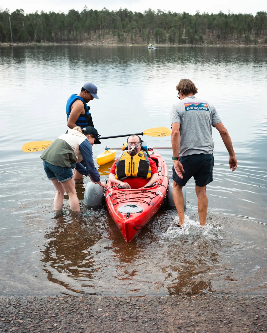 Ryan participating in adaptive kayaking with assistance at the edge of a lake.