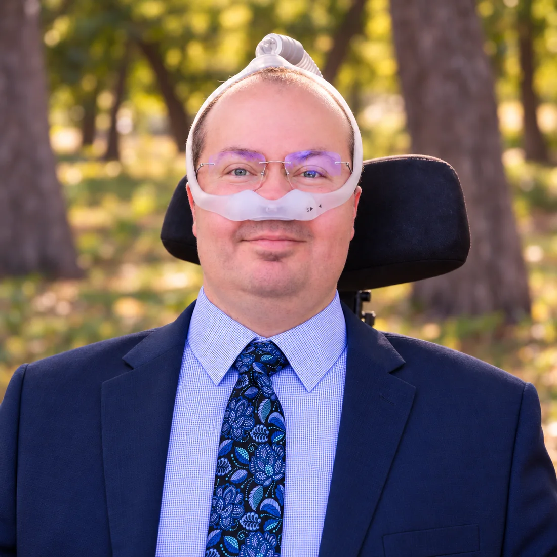 Ryan J. Russell, founder of FLDI, outdoors in a navy suit using a power wheelchair and respiratory support.